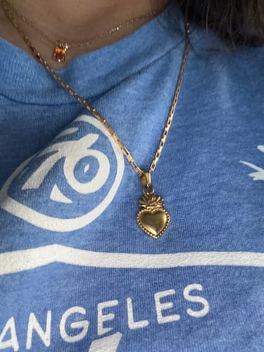Close-up of a reviewer wearing the gold Mini Amor Milagro Necklace against a blue t-shirt.