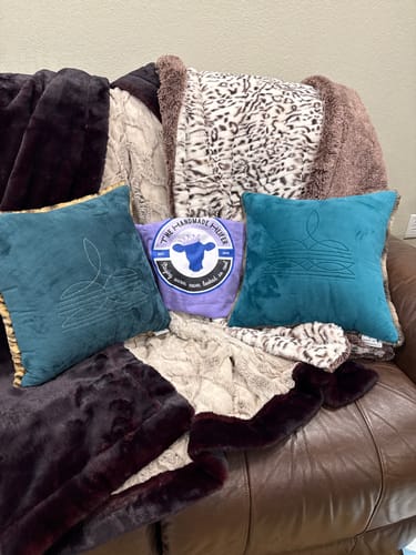Customer's two teal Boot Stitch Pillows shown on a leather sofa with other plush pillows and blankets.
