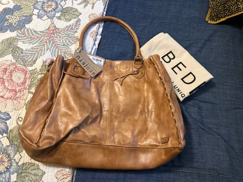 Customer's light brown Rockaway BedStu Purse sitting on a bed with its tag still on and the dust bag beside it.
