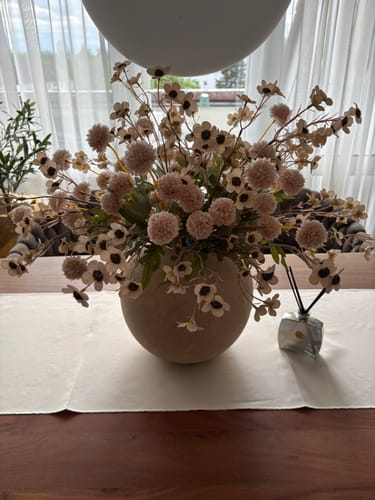 Recensent toont het Wax Arrangement - Crème in een ronde, beige vaas op een houten tafel.