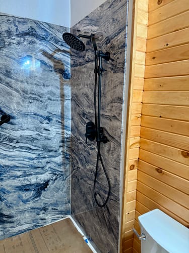 Reviewer's matte black 2-Handle Shower System installed against a dark, marbled shower wall in a wood-paneled bathroom.