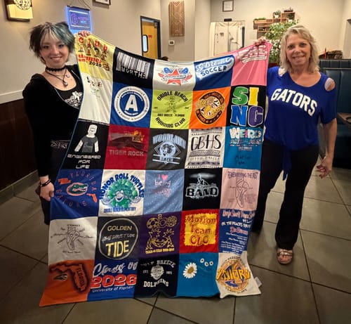 Customer and another person holding a large Project Repat T-Shirt Quilt made of many colorful shirts in a restaurant.