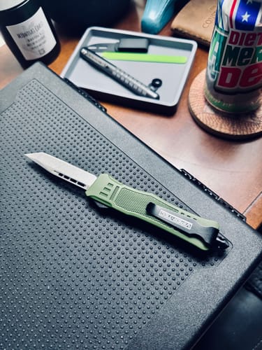 Reviewer's Small Green Phantom OTF knife with the tanto blade deployed, resting on a textured surface.