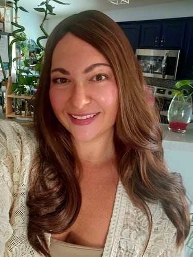 Reviewer smiling while wearing the long, brunette Cappuccino Blend Silk Topper in a well-lit kitchen.