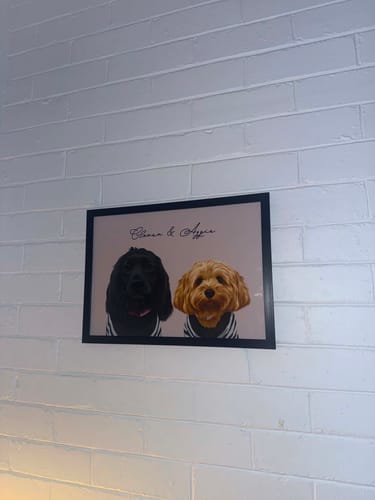 Customer's framed Two Pet Portrait of a black dog and a golden dog, hanging on a white brick wall.