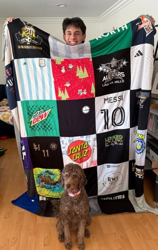 Customer holding a large Premium T Shirt Quilt made from various colorful shirts, with a dog sitting in front.