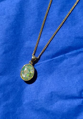 Customer's gold-plated Petite Oval Pendant with a greenish iridescent fill on a matching chain, resting on a blue surface.