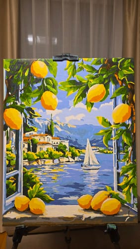 Ein Kunde zeigt das fertige „Malen nach Zahlen – Lago & Zitronen“-Bild auf einer Staffelei, das eine helle Seelandschaft darstellt.