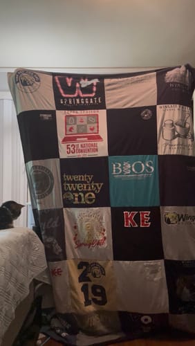 Customer holding up a huge T-shirt quilt made from many shirts in a bedroom, with a cat sitting on the bed looking on.