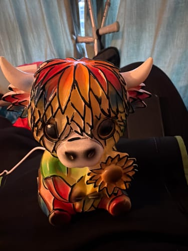 Reviewer's colorful Highland Cow LED Night Light sitting in a room. The cute resin lamp is holding a sunflower.