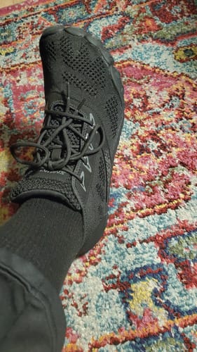 Customer showing their foot in a black Smart I Pro - Barefoot Shoe, viewed from above on a patterned rug.