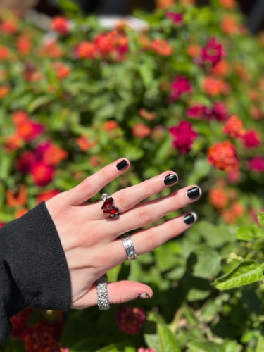 A reviewer's hand wearing the Promise Ring with red coffin stones on the index finger in front of flowers.