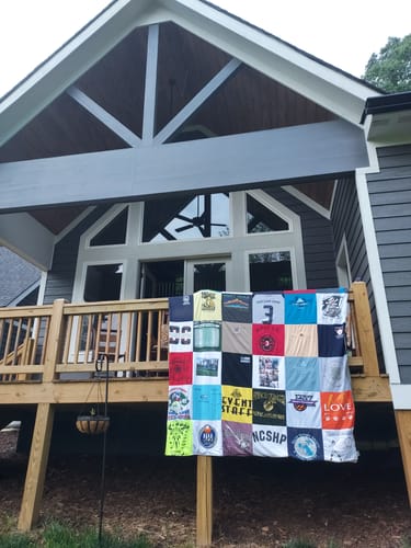 Customer's custom Project Repat T-Shirt Quilt displayed on a stand in front of their new home's deck.