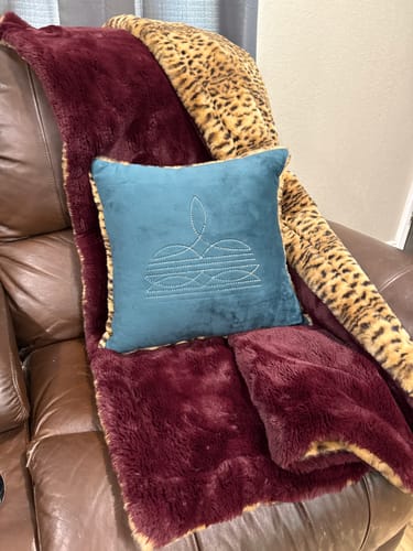 Customer's spruce Boot Stitch Pillow styled on a couch with a burgundy and leopard print blanket.