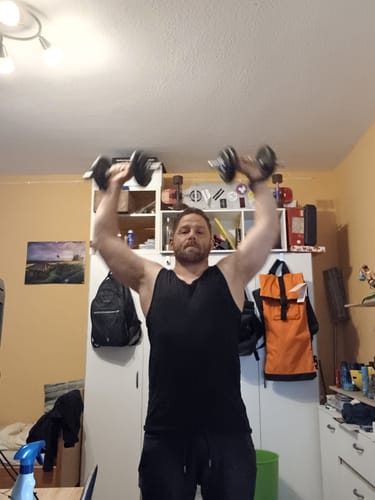 Reviewer working out at home, lifting two dumbbells overhead in a room.