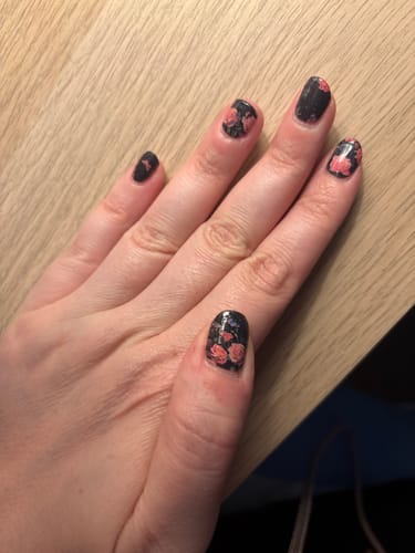 Hand einer Kundin, die die dunklen Midnight Garden Nagelfolien mit einem rosafarbenen Rosenmuster auf einer Holzoberfläche zeigt.
