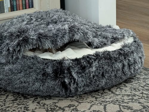 Customer's dog barely visible, snuggled deep inside the fluffy charcoal Pet Cave Dog Bed on a patterned rug.