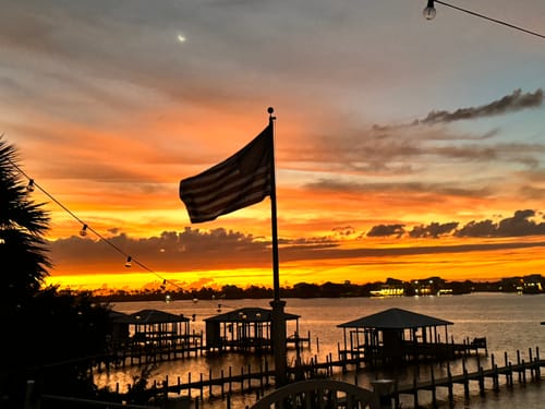 Customer's Ultra Heavy Duty American Flag silhouetted on a flagpole against a vibrant sunset over the water.