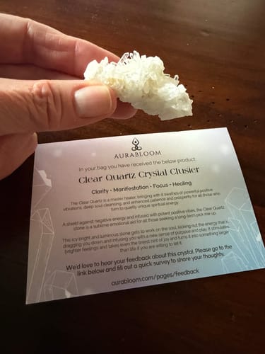 Reviewer holding a small Clear Quartz Crystal Cluster over its descriptive card on a wooden table.