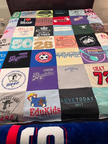 Customer's Project Repat T-Shirt Quilt spread on a bed, showing many colorful squares with various graphics and text.
