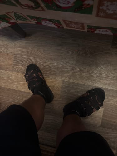 A reviewer's feet, viewed from above, wearing black Mystery DracoSlides with a printed pattern and black socks indoors.