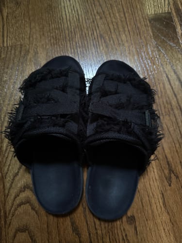 Reviewer's pair of black slides with frayed, textured straps on a wood floor.