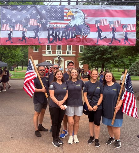 Reviewers in gray 250 Years of Heroes shirts posing under a 'Because of the Brave 5K' banner.