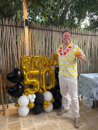 Customer posing with a large balloon centerpiece featuring gold foil balloons that spell "BRAD 50" for a party.