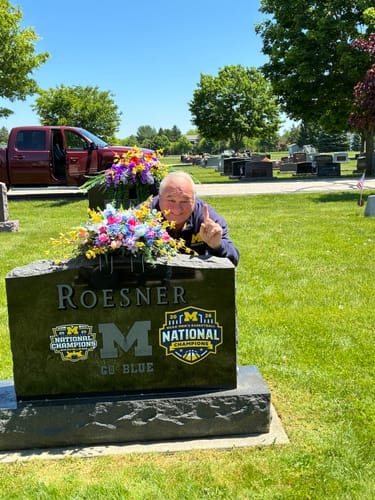 Customer posing behind a gravestone displaying the Michigan Wolverines 2026 Basketball Champions plaque and another.