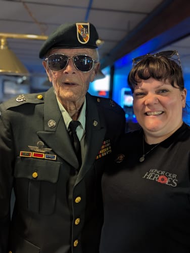 Reviewer wearing her black 'The Promise We Keep' tee, standing proudly with a decorated veteran.