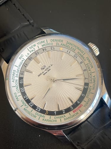 Reviewer's close-up of the silver World Time GMT watch, showing its textured white dial and black leather strap.