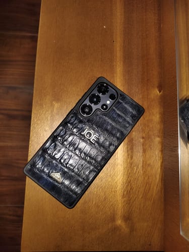 Reseñador mostrando una funda S25 Ultra Croc azul marino personalizada con el nombre 'JOE' sobre una mesa de madera.