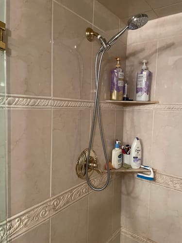 Reviewer's silver handheld shower head installed in a tiled shower stall with gold fixtures.