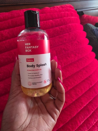 Customer holding a clear bottle of Yummy Body Splash body oil against a red textured background.