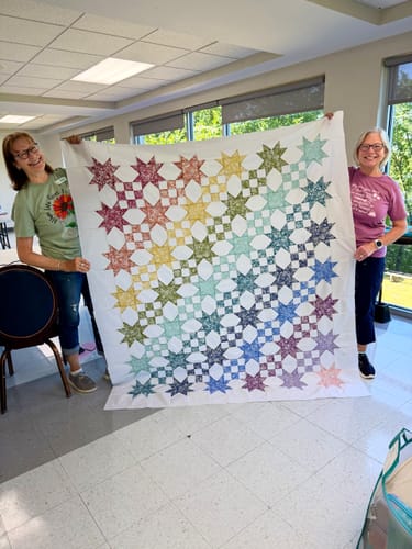 Two reviewers holding up the finished Star Studded Quilt top. The large quilt has a colorful, repeating star pattern.