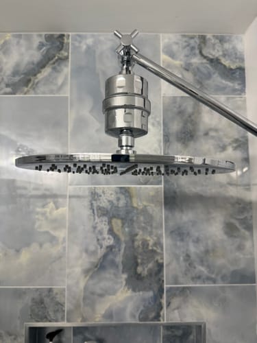 Customer's chrome Shower Head Filter installed on a large rain shower head against a marbled tile wall.