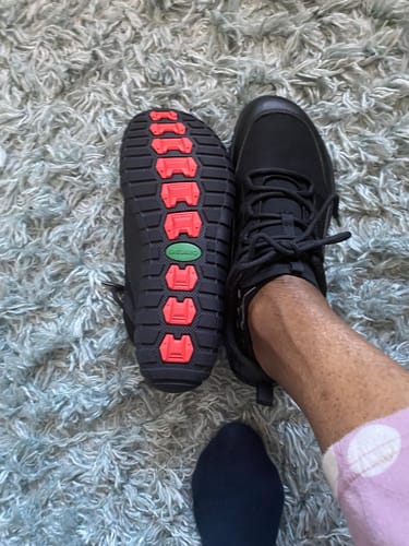 Reviewer showing the sole of one black Forestep Ⅰ Barefoot Shoe, with its red tread, next to the other shoe on their foot.