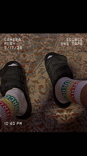 A reviewer's feet wearing black slides with textured straps over white socks on a patterned carpet.