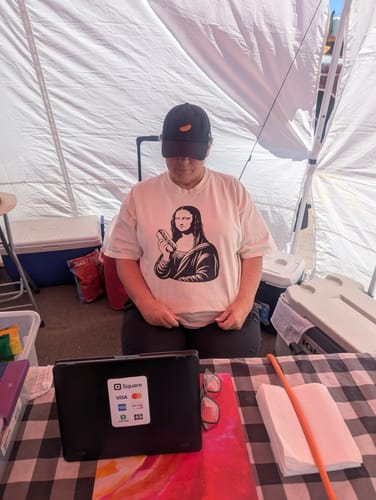 Customer wearing a white Mona Wiener — Premium Tee while sitting at a market stall.