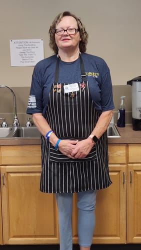 Reviewer standing in a kitchen wearing the blue 250 Years of Heroes t-shirt under a striped apron.
