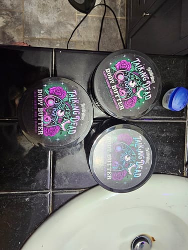 Customer's three tubs of Talking Dead Body Butter arranged on a black bathroom counter next to a sink.