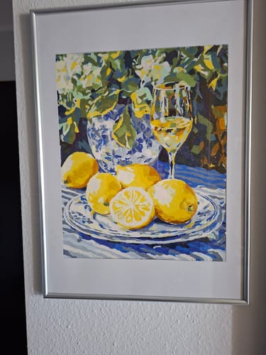 Ein Kunde zeigt das fertige "Malen nach Zahlen - Zitrone und Wein" Bild, das in einem silbernen Rahmen an einer weißen Wand hängt.
