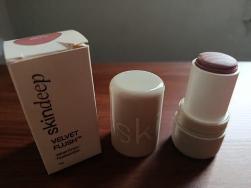 Customer's open VELVET FLUSH Lip+Cheek Blush stick, showing its mauve color next to its box on a wooden table.