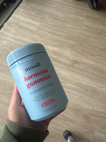 Customer holding a blue container of Moodi Hormone Gummies in the Strawberry flavor against a wood-look floor.