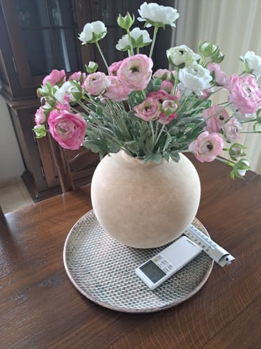 Recensent toont het Ranonkel Arrangement met roze en witte bloemen in een ronde, beige vaas op een tafel.