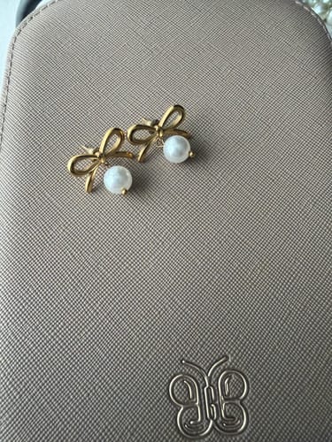 Customer's Laila Bow Pearl Earrings, showing the gold bow design and dangling pearls on a textured surface.