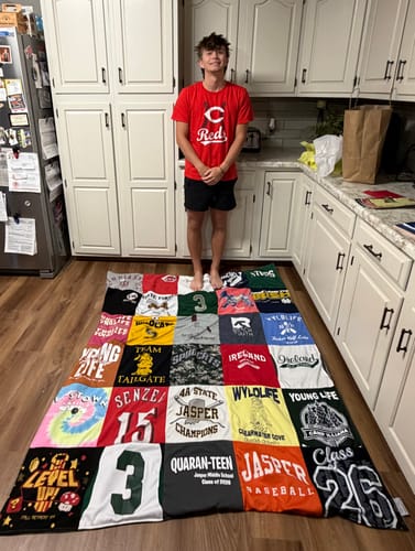 Reviewer showing a large, colorful Project Repat T-Shirt Quilt made from many shirts, spread out on a kitchen floor.