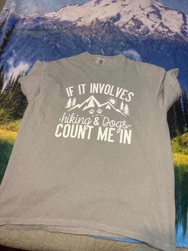 Customer's gray 'If It Involves Hiking And Dogs' t-shirt laid out flat on a mountain-print blanket.