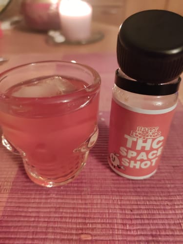 Kunde zeigt den THC Space Shot Orange in einem Totenkopfglas mit Eis, daneben die Originalflasche.