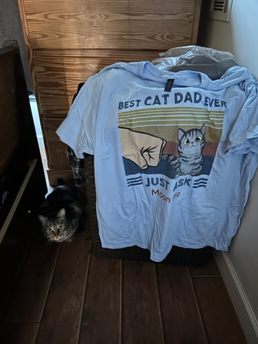 Customer's personalized "Best Cat Dad Ever" shirt in light blue, with their cat, Moon, sitting on the floor beside it.
